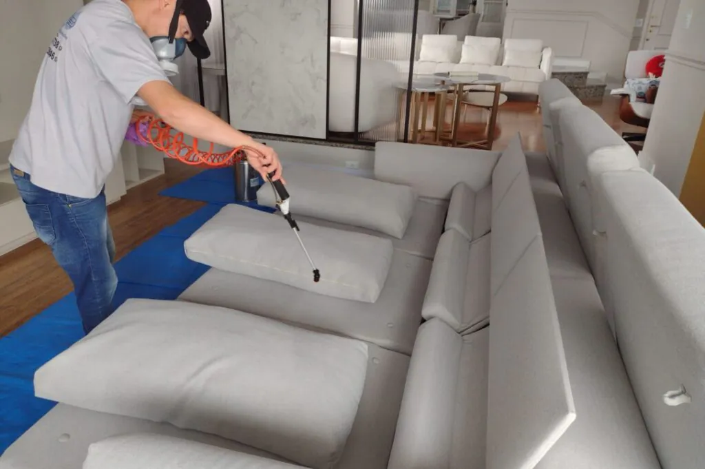 Profissional aplicando impermeabilizante em um sofá em ambiente claro e moderno, utilizando equipamento de pulverização com proteção adequada.